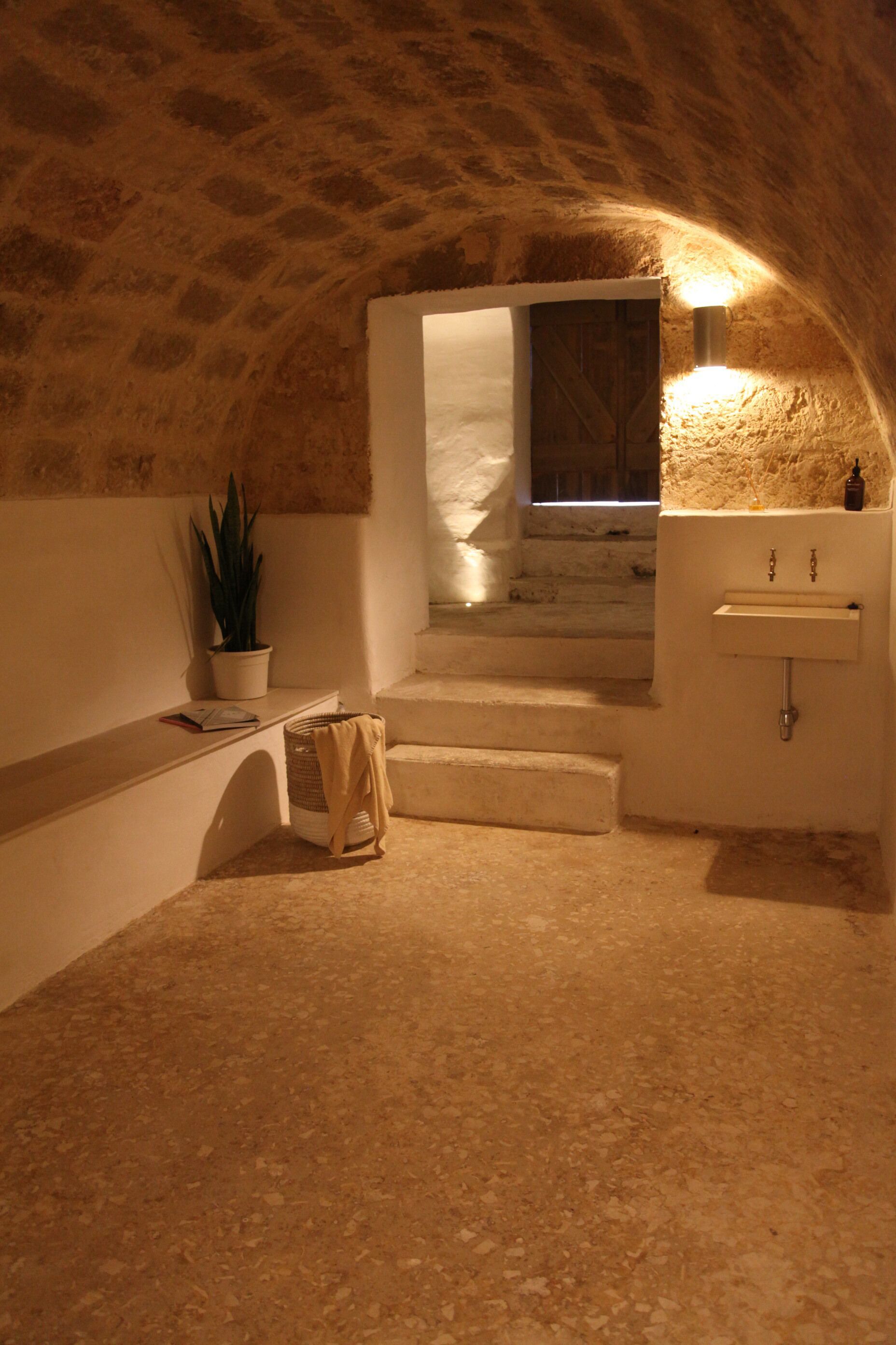 A stone-walled bathroom with a sink, a potted plant, and a staircase leading to a window.