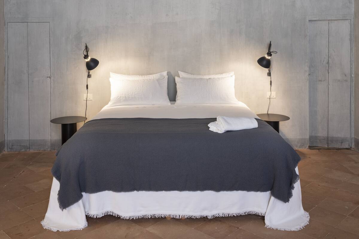 Et soveværelse med en seng, to sengeborde med lamper, et gråt og hvidt tæppe samt et mønstret loft.