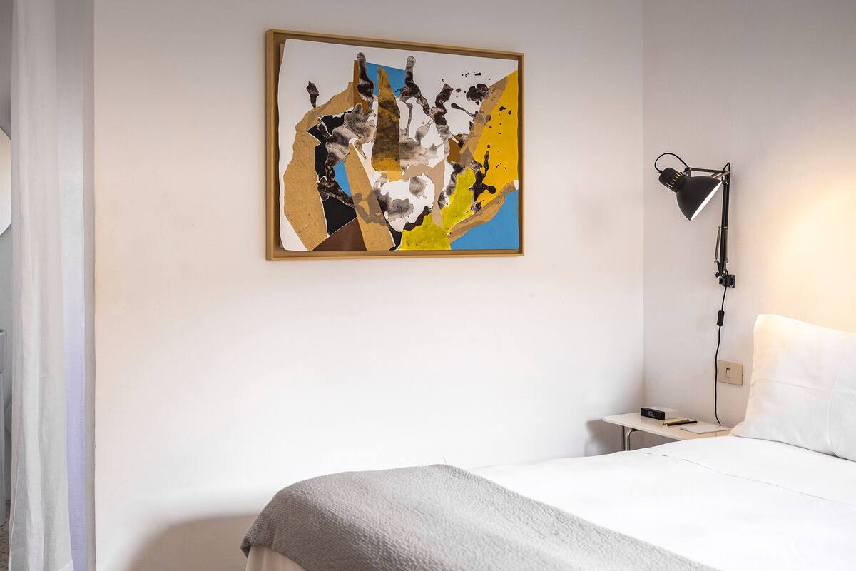 Et hotelværelse med en seng, et natbord med en lampe, et abstrakt maleri på væggen, et rundt spejl og et toilet.