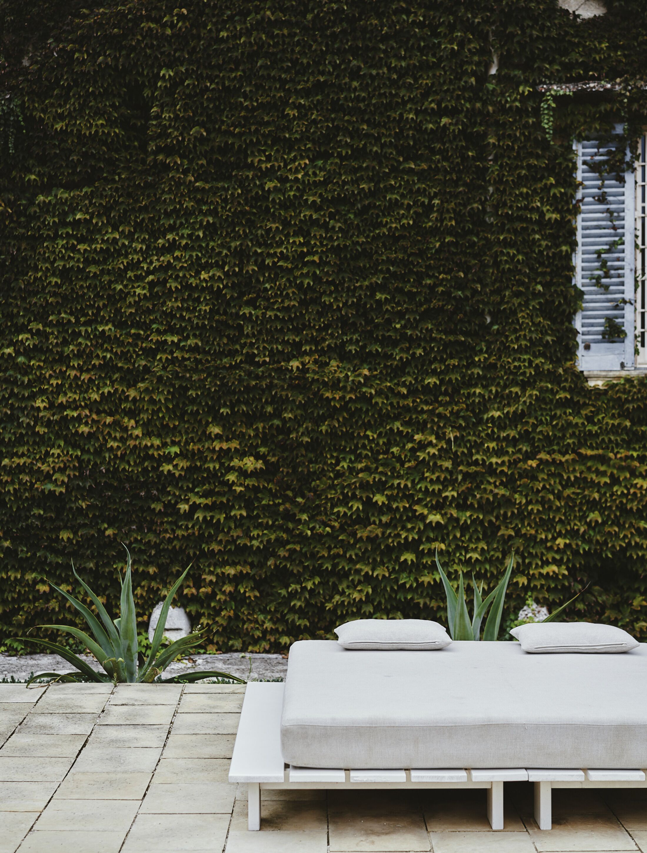 A white outdoor seating area with a bench and cushions, surrounded by a hedge wall and a window with shutters.