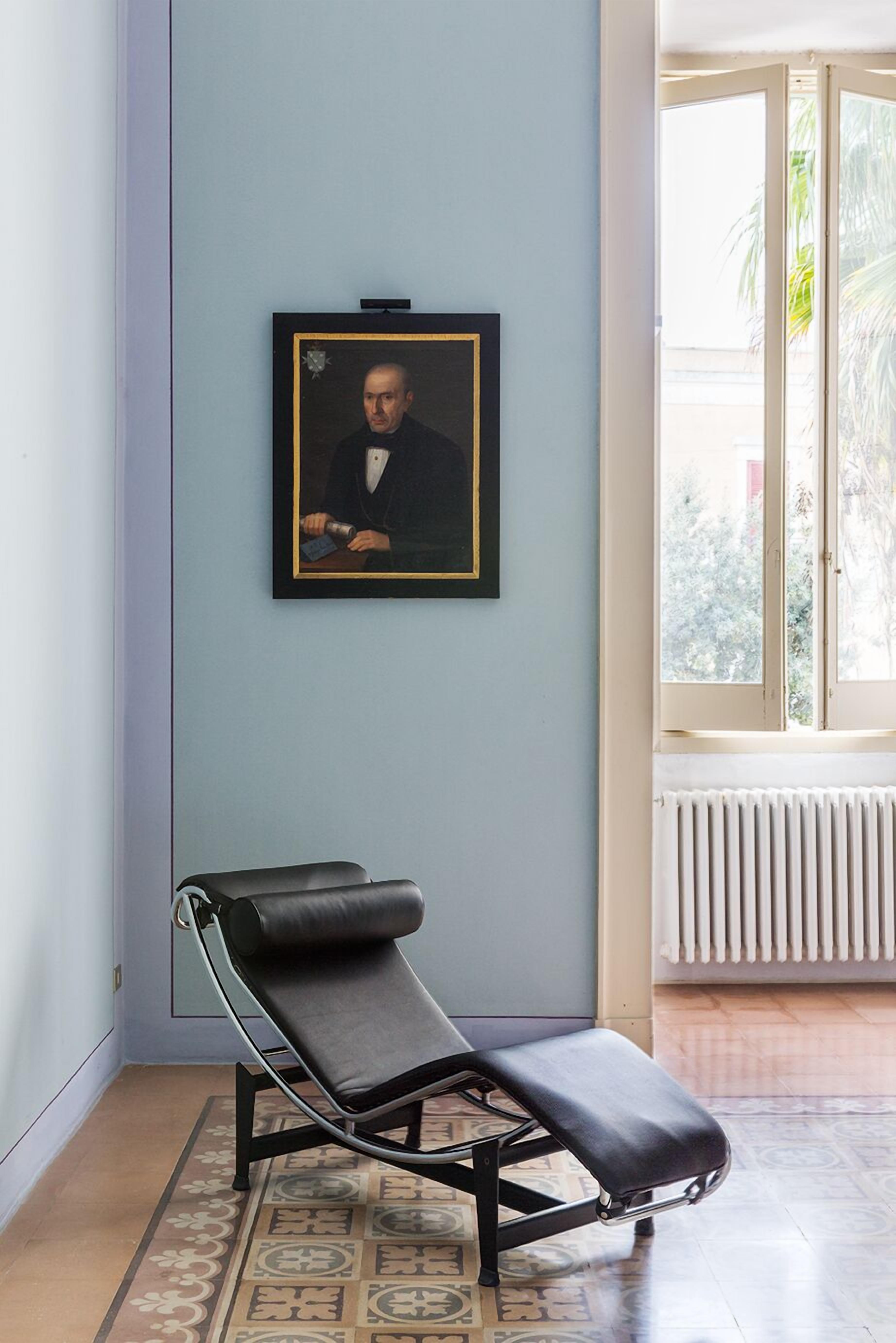 A room with a framed portrait on the wall, a black lounge chair, a patterned rug, and a window with a view of greenery.