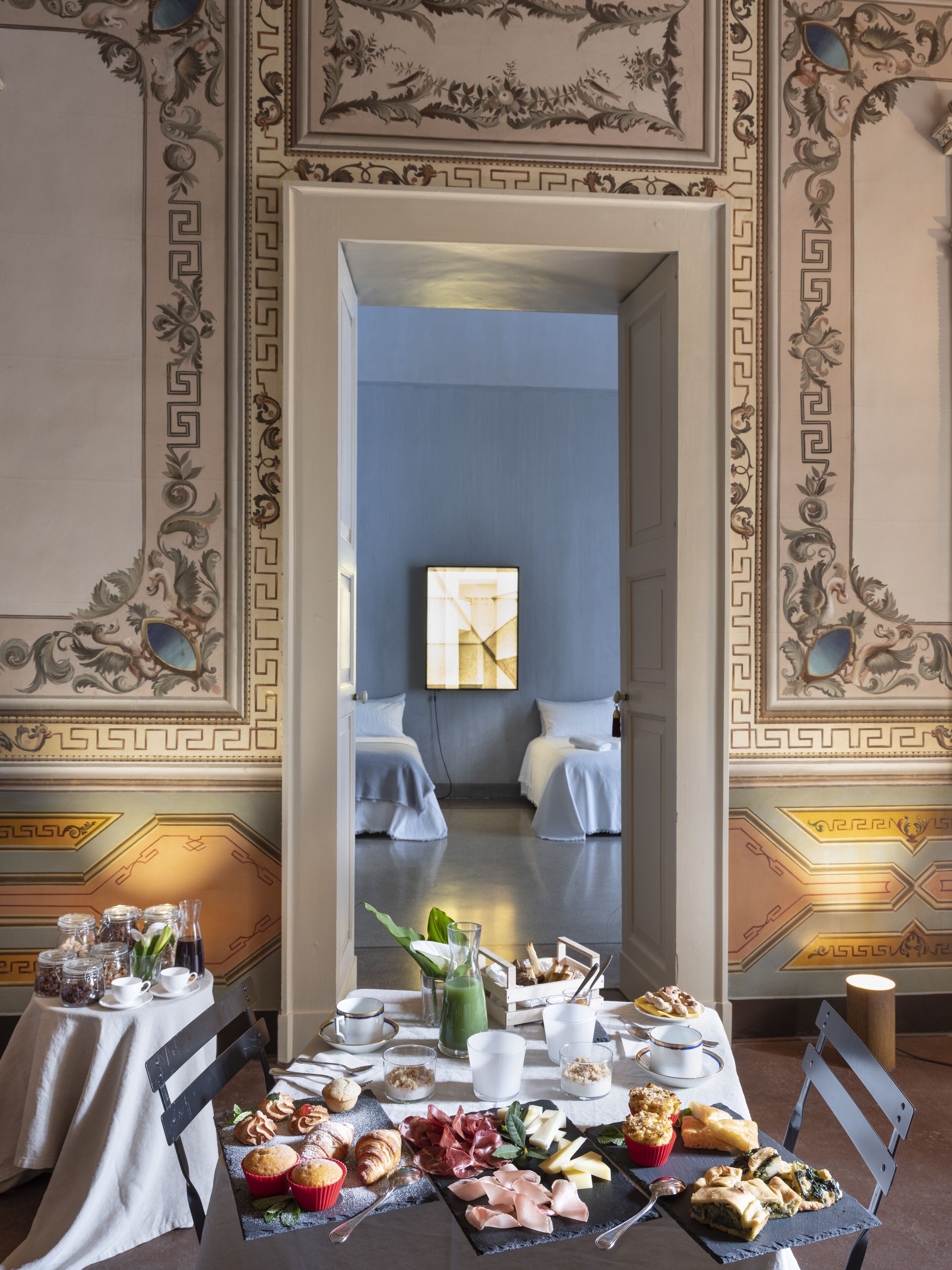 Zona pranzo con tavolo apparecchiato per la colazione, con pasticcini, formaggi e salumi. La stanza presenta decorazioni murali elaborate e vista su una camera da letto con un letto e un quadro appeso.