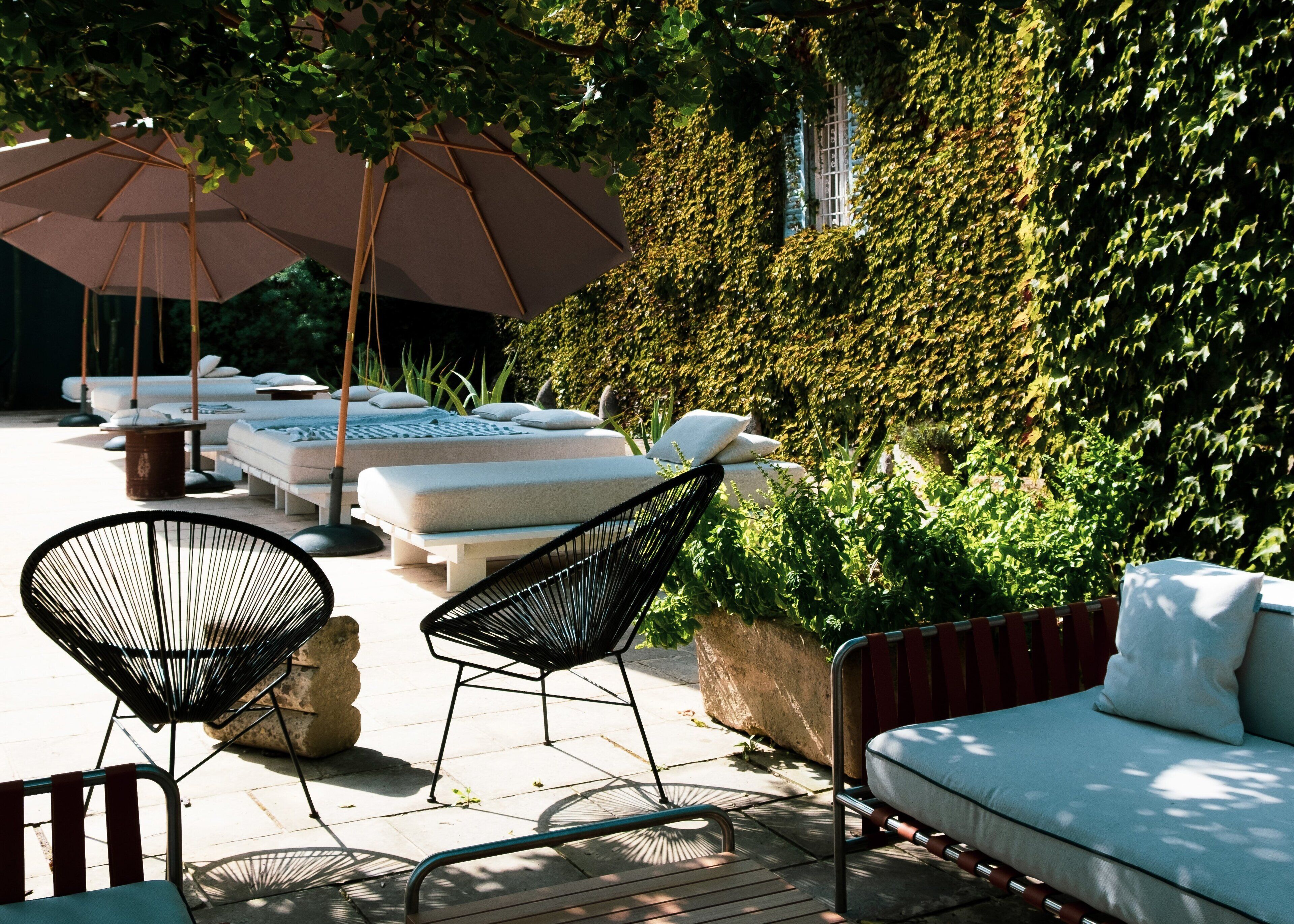 An outdoor seating area with lounge chairs and a large umbrella, surrounded by lush greenery and a wall covered in ivy.