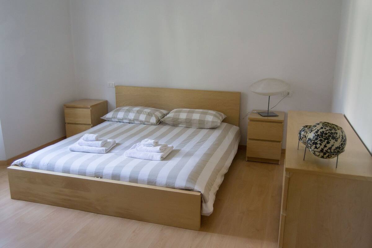 Un lit bien fait, avec une literie à rayures et deux oreillers, une table de chevet avec une lampe, et un vase décoratif posé sur une commode en bois.