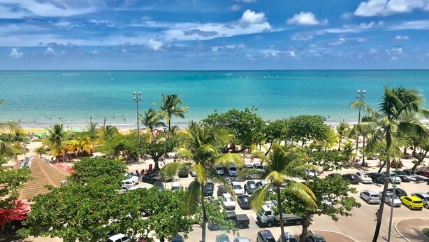 A beach view with a parking area, palm trees, and a clear sky. - Happy Hotel Pajuçara (Maceió)