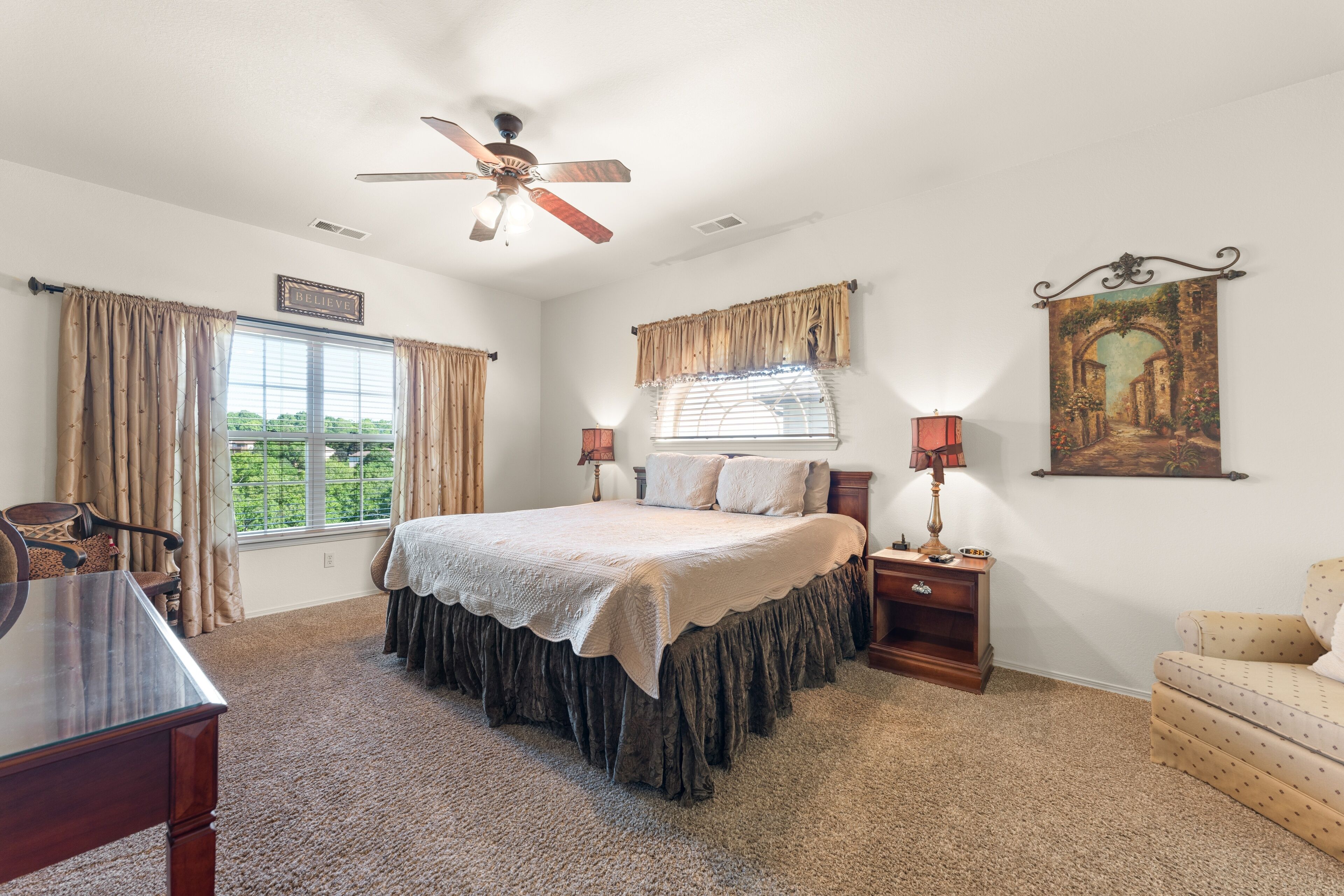 A bedroom with a large bed, two bedside tables, a chair, a desk, a ceiling fan, and a window with curtains.