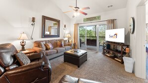 A living room with a leather sofa, a coffee table, a TV on a stand, a ceiling fan, and a view of the outdoors through a sliding glass door.