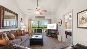 A living room with a sofa, ottoman, ceiling fan, TV, and a view through glass doors.