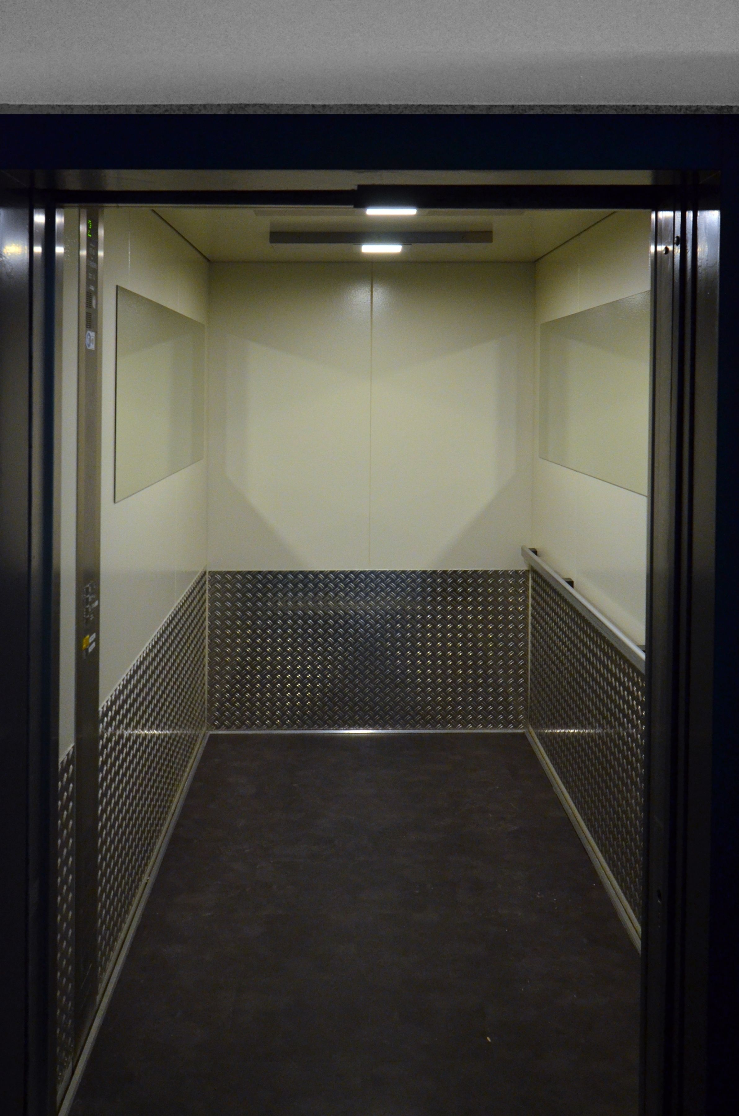 An elevator with a metallic floor and walls, featuring a control panel on the left and a door on the right.