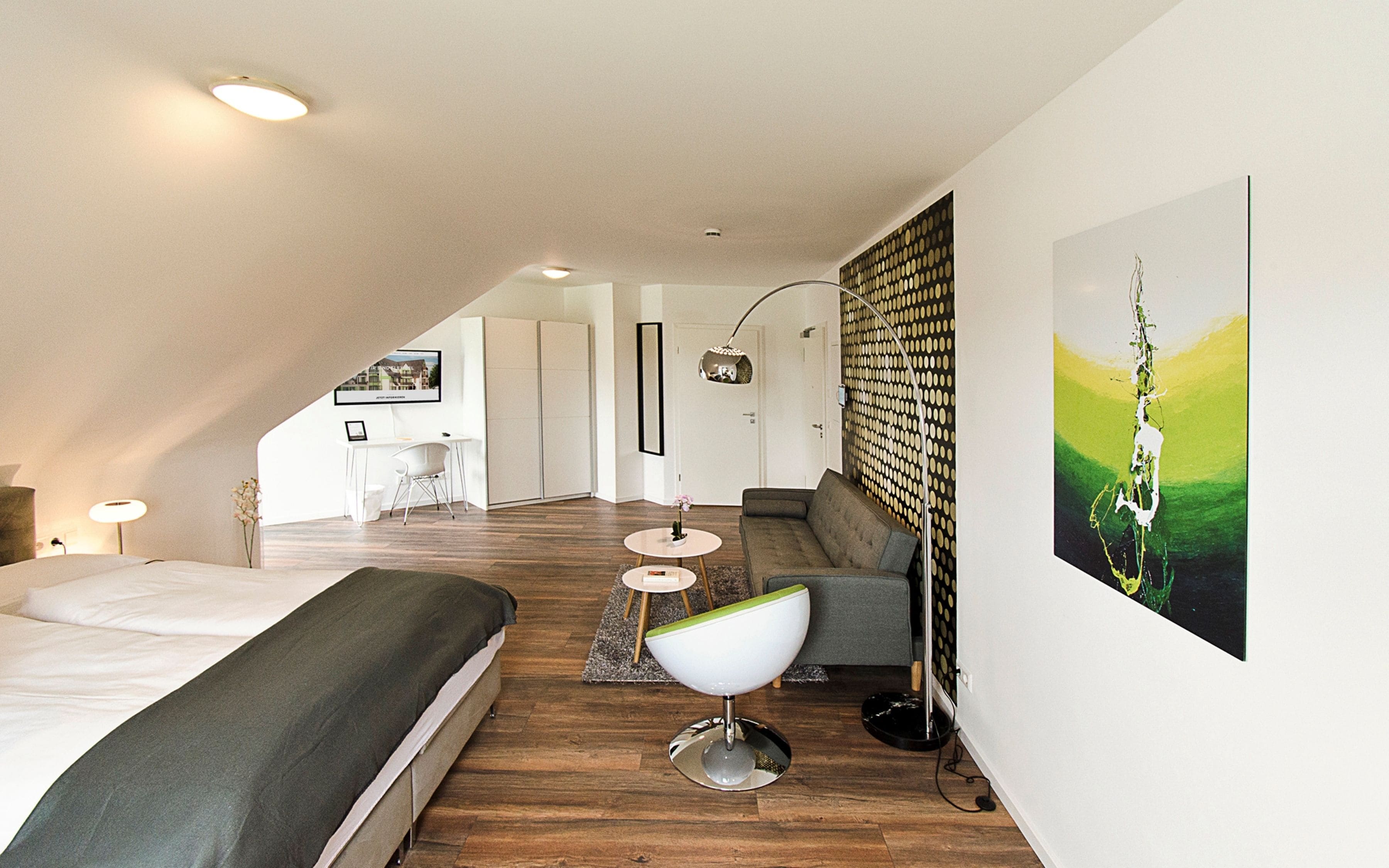 A modern bedroom with a large bed, a desk, and a sofa. There is a painting on the wall and a lamp on the desk.