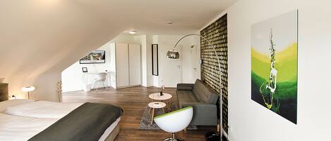 A modern bedroom with a large bed, a desk, and a sofa. There is a painting on the wall and a lamp on the desk.