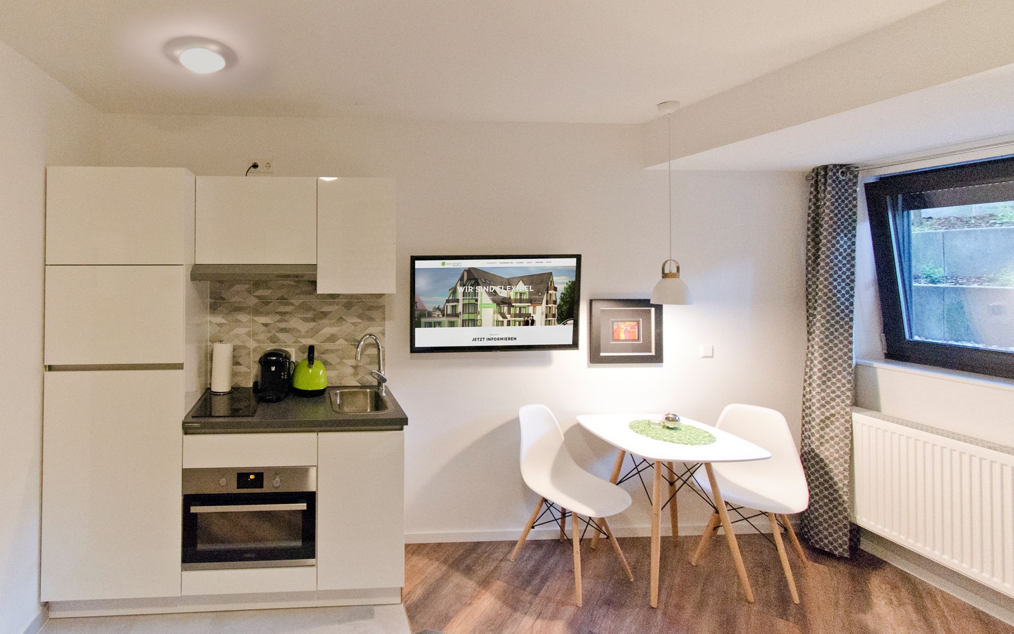 A compact kitchen with a built-in oven, a coffee maker, and a sink. There is a dining area with two chairs and a small table. A framed picture and a digital display are mounted on the wall.
