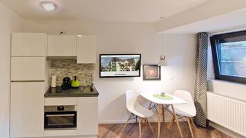 A compact kitchen with a built-in oven, a coffee maker, and a sink. There is a dining area with two chairs and a small table. A framed picture and a digital display are mounted on the wall.