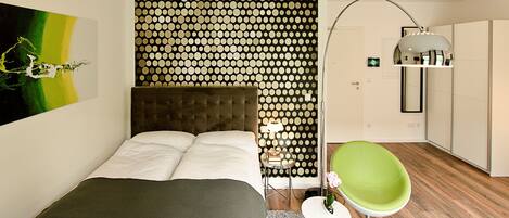 A modern bedroom with a polka dot wall, a tufted headboard, a green chair, and a bedside table with a lamp.