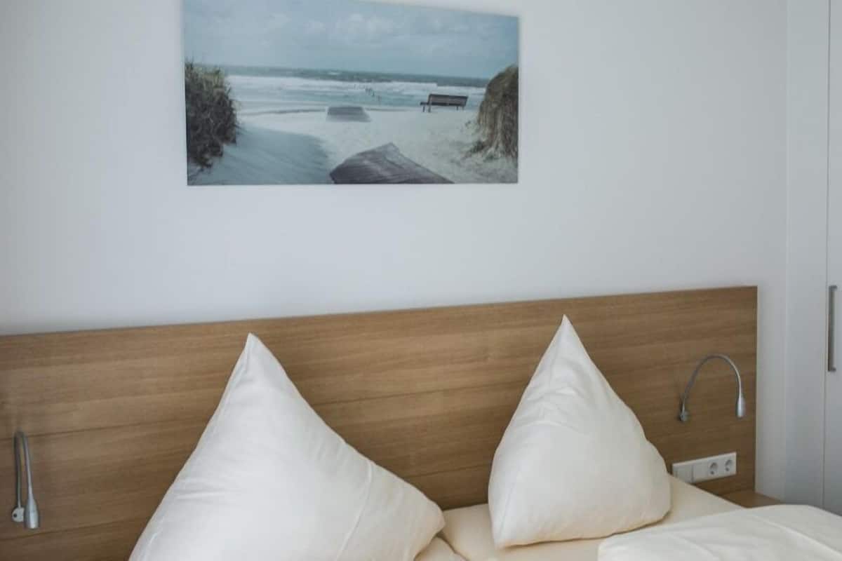 Ein Schlafzimmer mit einem hölzernen Kopfteil, zwei weißen Kissen, einem Nachttisch mit Lampe und einem Bild einer Strandlandschaft an der Wand.