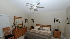 Quarto com cama, penteadeira, cadeira, mesas de cabeceira e ventoinha no teto.