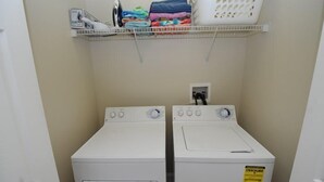 Uma lavanderia com duas máquinas de lavar de carregamento frontal brancas, uma cesta de roupa branca numa prateleira e uma parede branca.