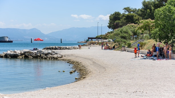 Une plage avec un rivage rocheux, un parasol rouge et des gens qui se détendent.