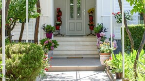 Une véranda à l’avant de la maison, avec une porte blanche, entourée de plantes en pot et d’arbres.