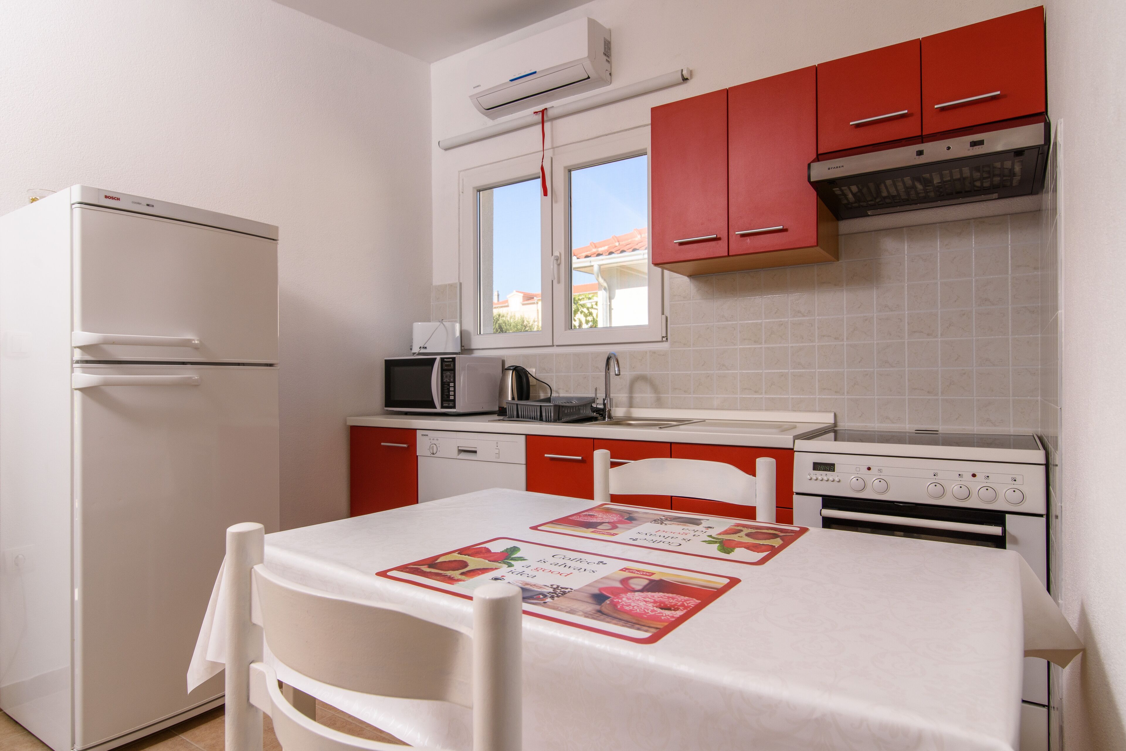 A compact kitchen with white appliances, red cabinets, and a window above the sink.