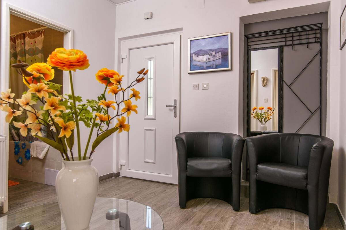 Une pièce moderne avec une table en verre, un vase blanc orné de fleurs, deux fauteuils en cuir noir, une porte et un tableau encadré accroché au mur.