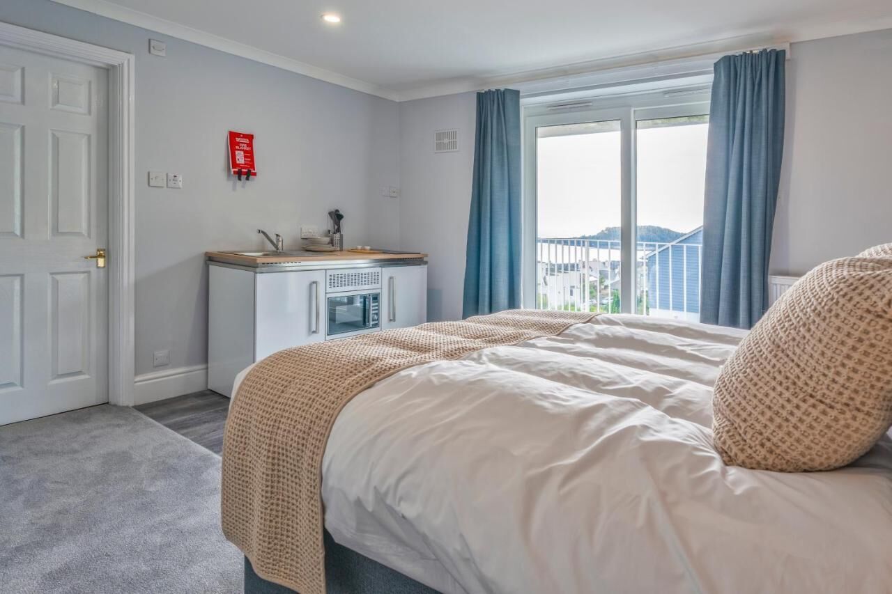 Une chambre d’hôtel avec un lit, une petite kitchenette, une fenêtre avec des rideaux et une vue sur les bâtiments environnants.