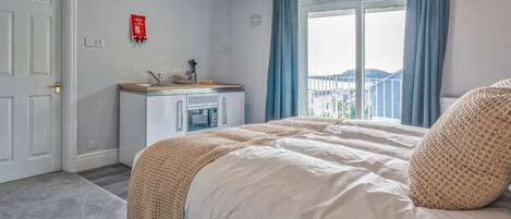 A hotel room with a bed, a small kitchenette, a window with curtains, and a view of buildings outside.