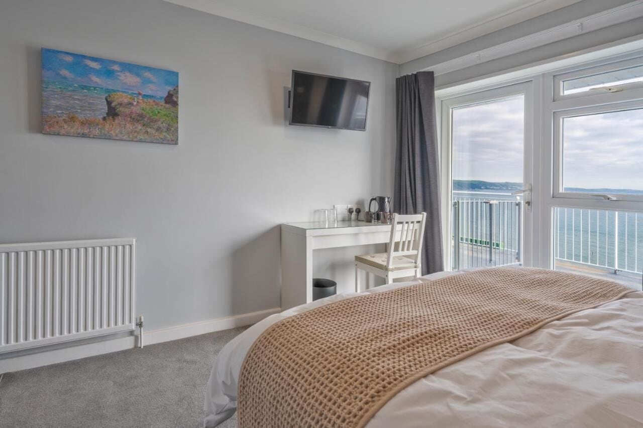 A bedroom with a bed, a desk, a chair, a TV, and a balcony with a view of the sea.