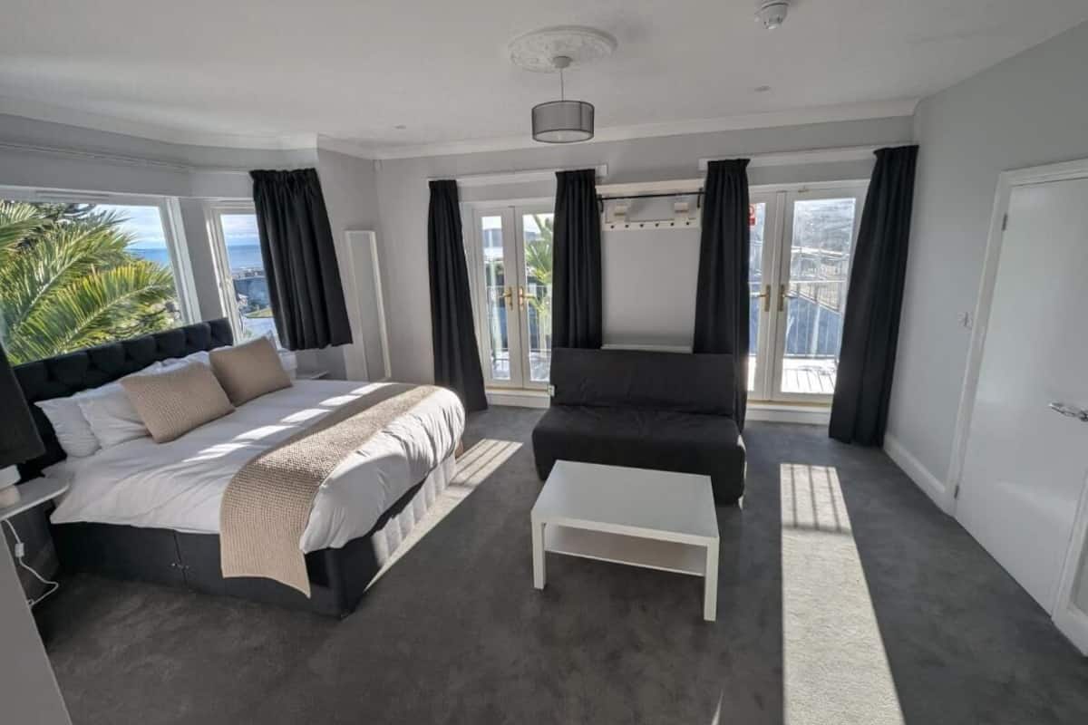 Quarto moderno com cama, poltrona preta, mesa de centro branca e janelas grandes com cortinas.