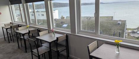 A dining area with tables and chairs, large windows with a view of the sea and buildings.