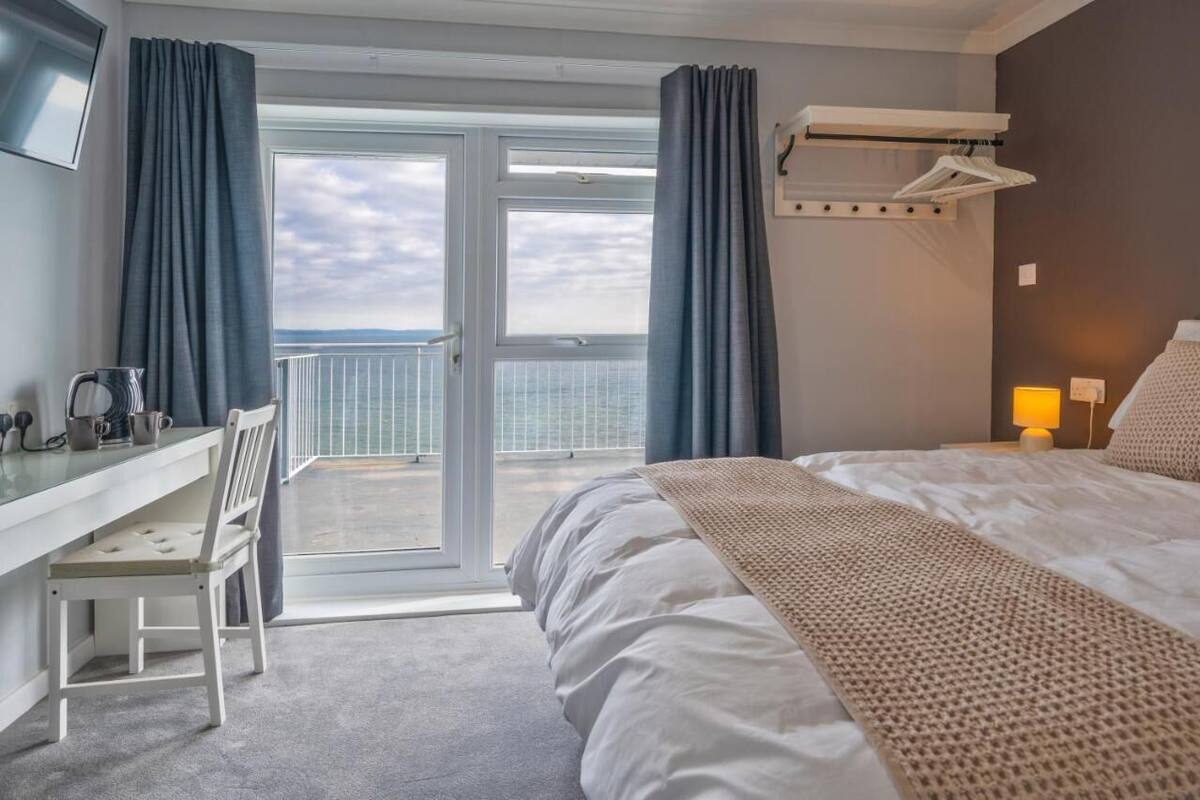 Quarto de hotel com cama, escrivaninha, cadeira, varanda com vista para o mar e um cabideiro.