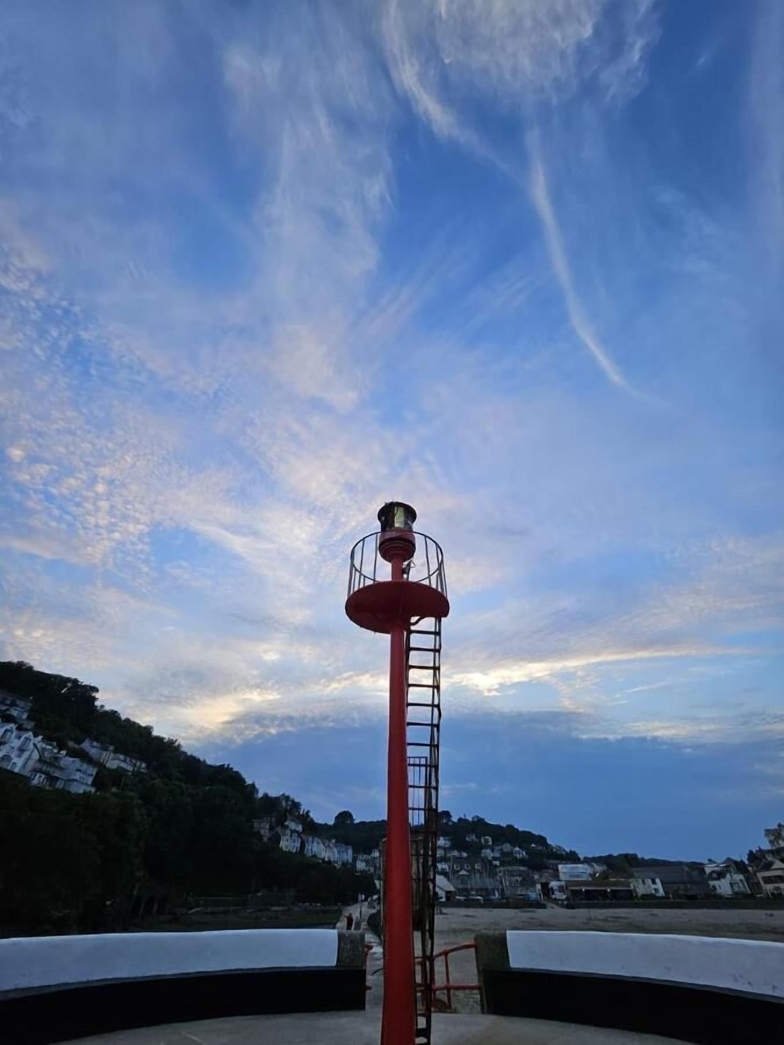 Un faro con una plataforma circular y barandilla roja, que domina una zona costera con edificios y colinas.
