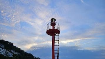 Un faro con una plataforma circular y barandilla roja, que domina una zona costera con edificios y colinas.