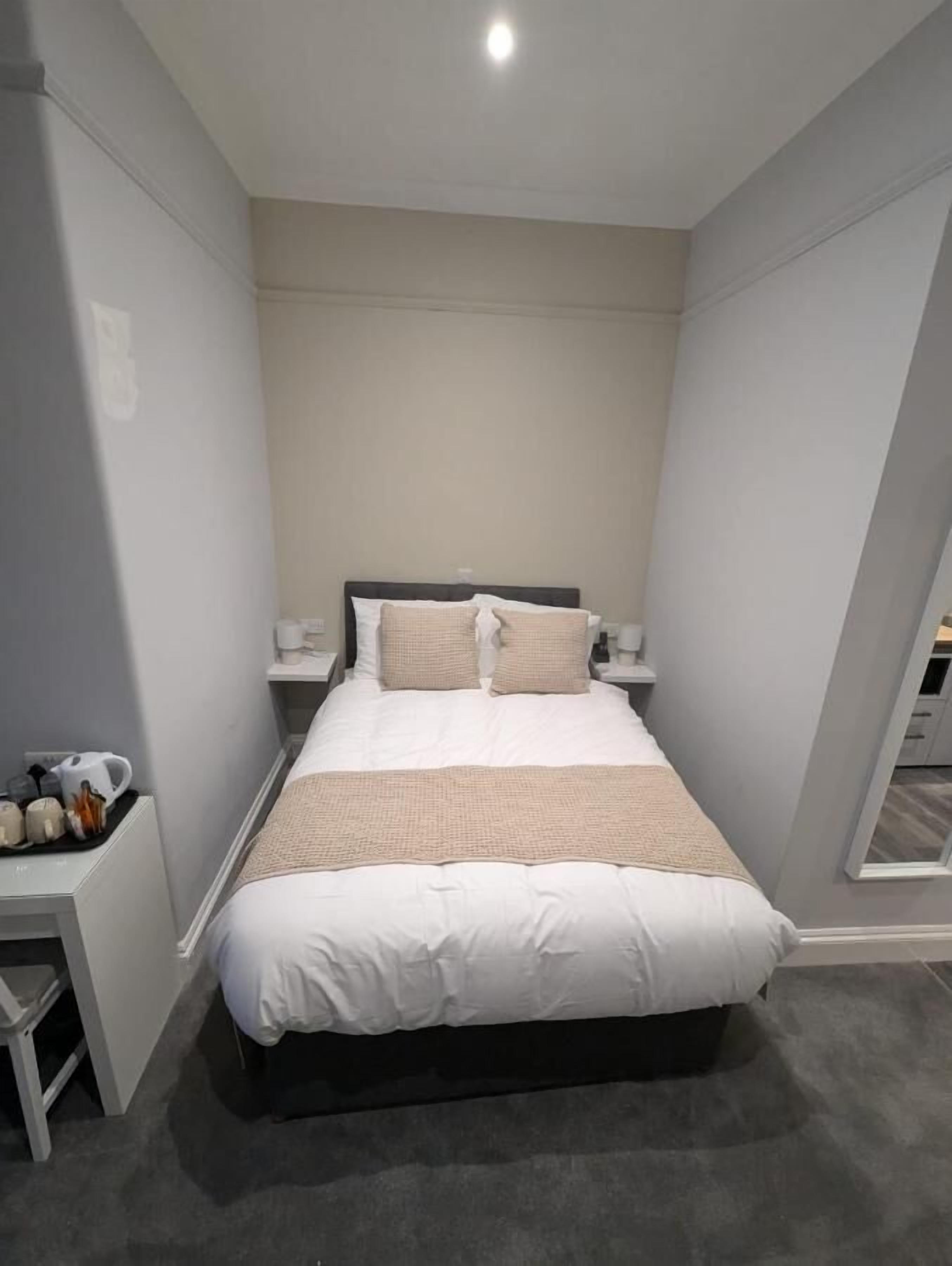 A modern bedroom with a large bed, a bedside table with a lamp, and a small table with a kettle and cups.