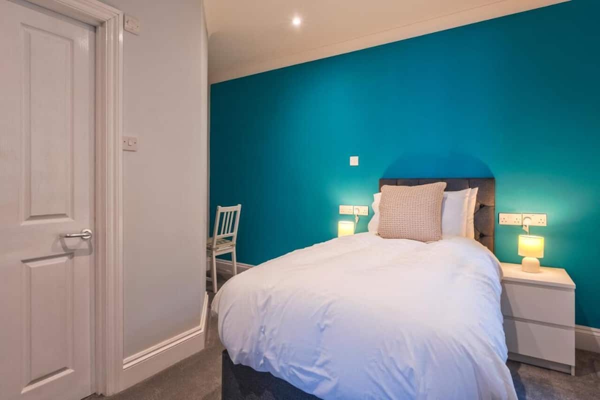 Quarto com parede verde-água, porta branca, cama com roupa de cama branca e mesa de cabeceira com abajur.