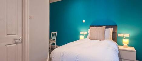 A bedroom with a teal wall, a white door, a bed with white bedding, and a nightstand with a lamp.