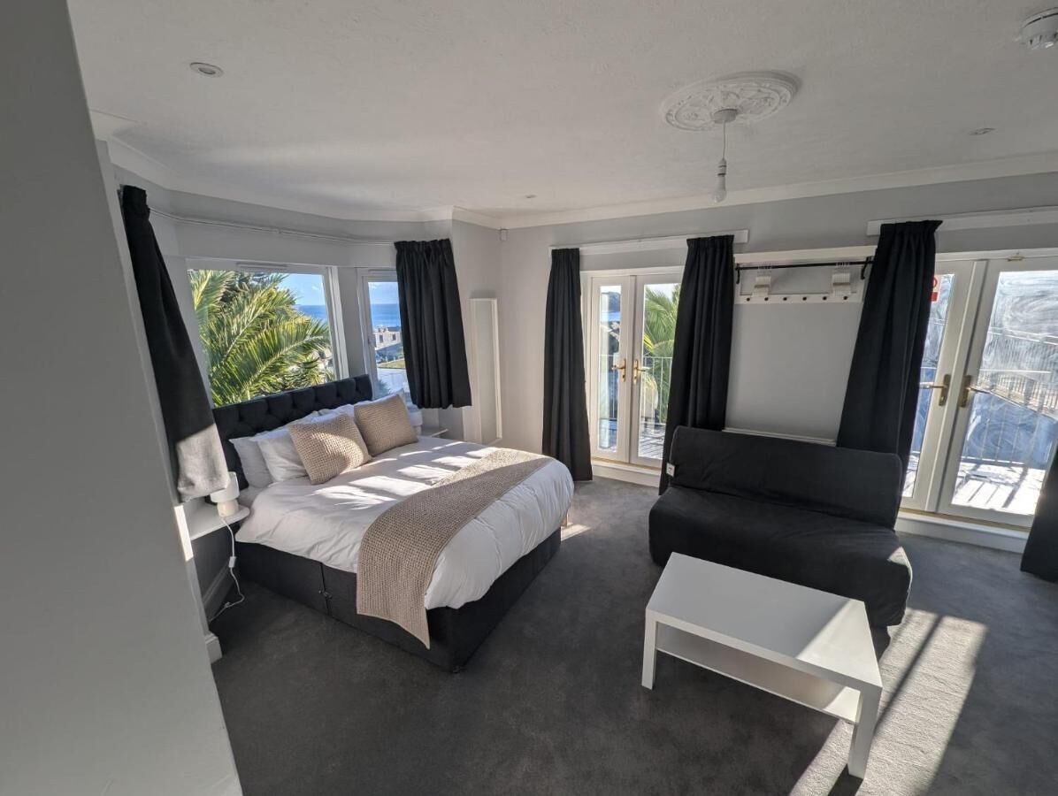 A modern bedroom with a large bed, a sofa, and a small table. There are large windows with curtains, and a view of palm trees and the ocean.