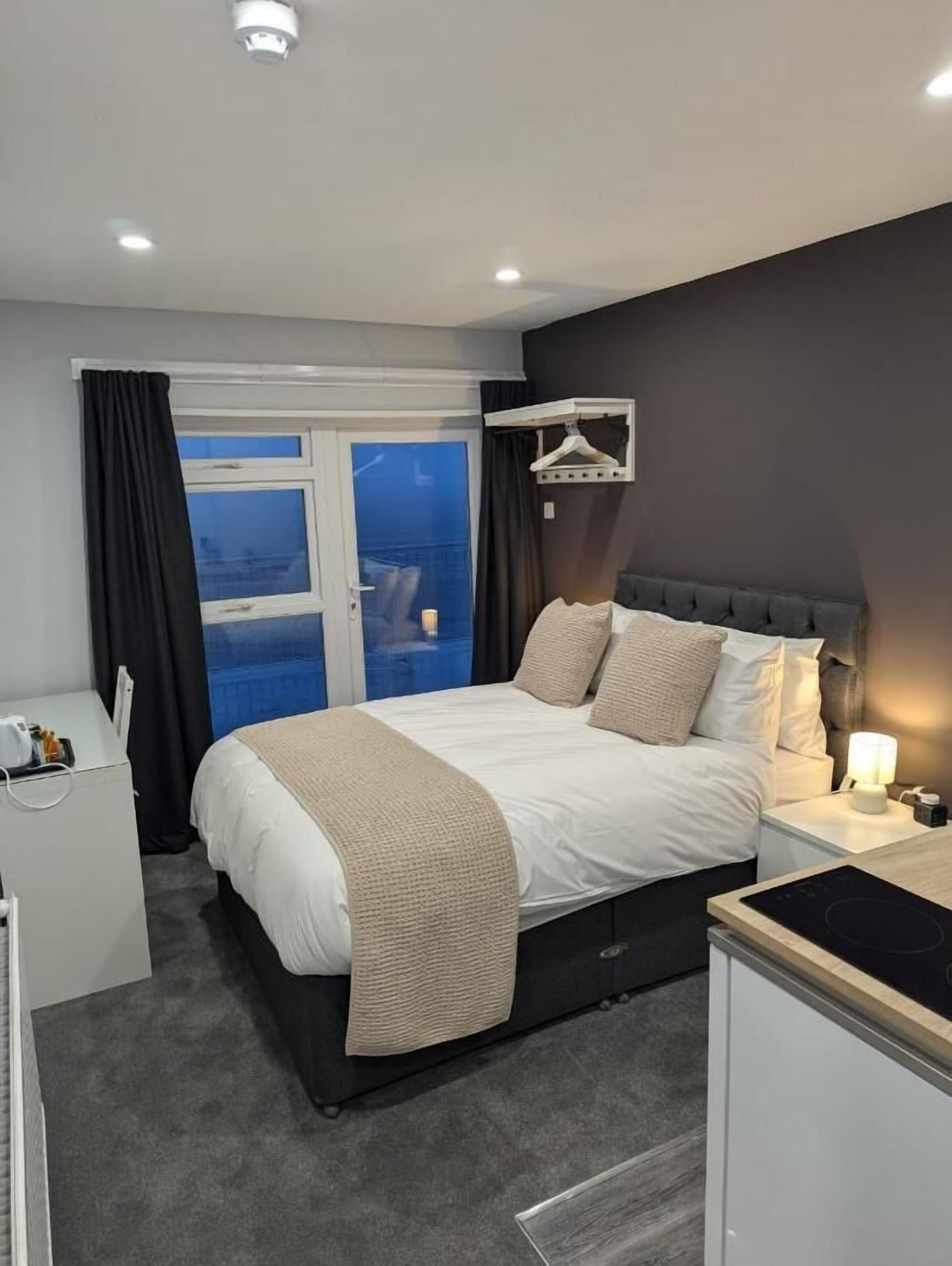 A modern hotel room with a large bed, a bedside table with a lamp, a small desk, and a view of the city through a large window.