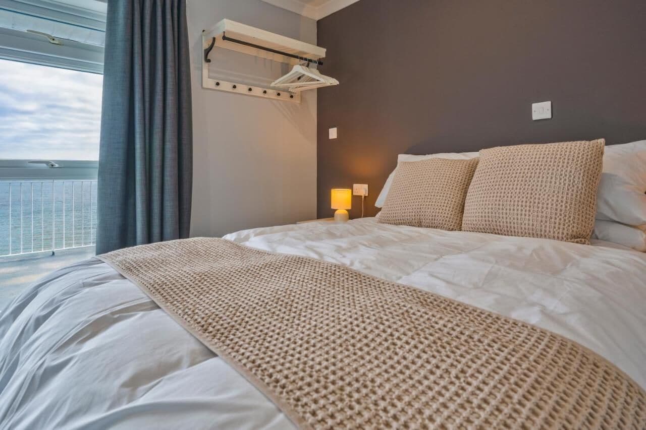 Habitación de hotel con una cama grande, un perchero, una mesita de noche con lámpara y vista al mar a través de una ventana.
