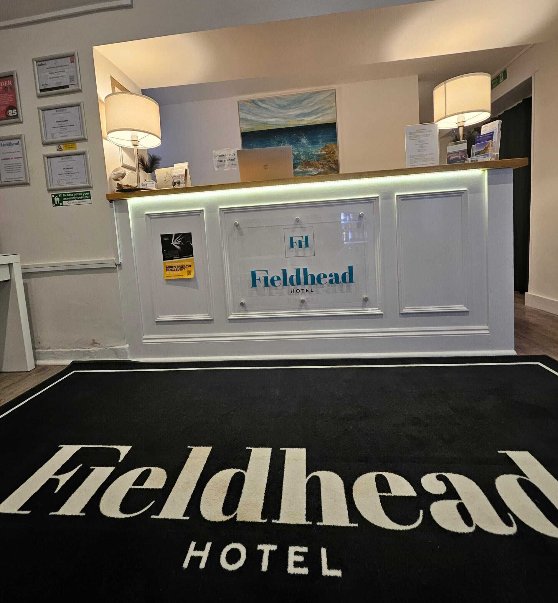 Mostrador de recepción en el Fieldhead Hotel con una alfombra negra y texto blanco, certificados enmarcados en la pared y un cuadro sobre el mostrador.
