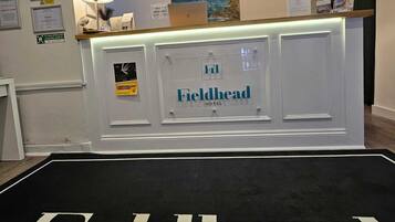 Mostrador de recepción en el Fieldhead Hotel con una alfombra negra y texto blanco, certificados enmarcados en la pared y un cuadro sobre el mostrador.