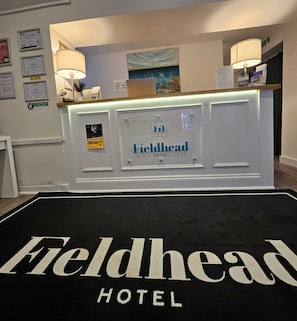 A reception desk at Fieldhead Hotel with a black carpet with white text, framed certificates on the wall, and a painting above the desk. - Fieldhead Hotel (Looe)