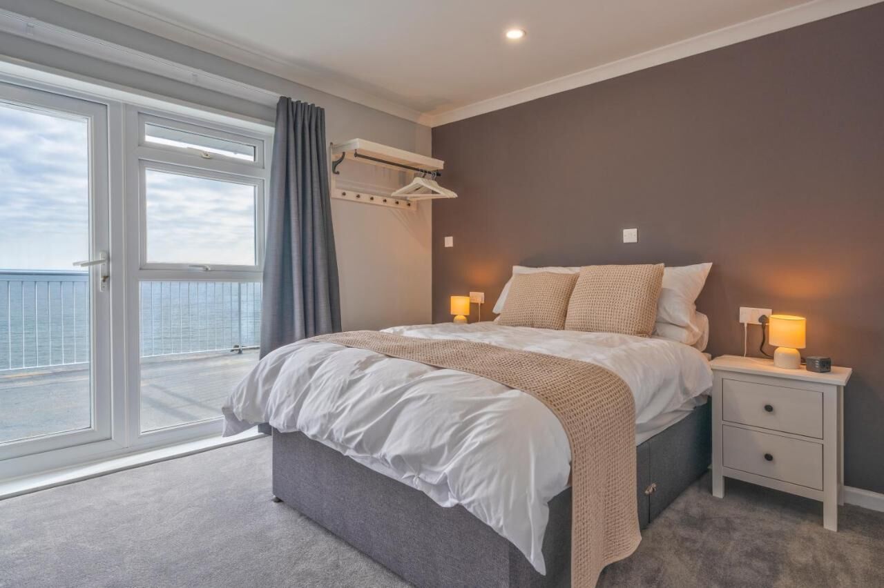 Ein Schlafzimmer mit einem großen Bett, einem Nachttisch, einer Lampe, einem Fenster mit Vorhängen und einem Balkon mit Meerblick.