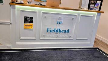 A hotel reception desk with a framed painting above it, a sign indicating a food allergy or intolerance policy, and a welcome mat at the entrance.
