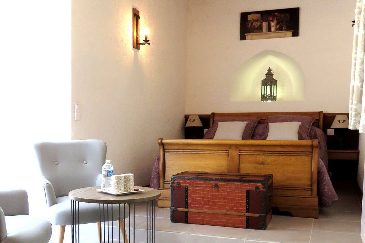 Quarto com cama de madeira, uma mesa redonda pequena com uma garrafa de água, uma cadeira e uma mala antiga.