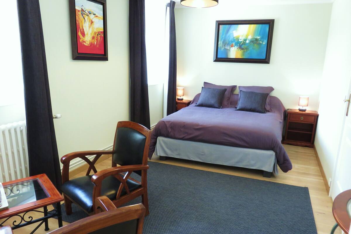 Quarto com cama, duas mesas de cabeceira, uma cadeira, uma mesa e duas pinturas na parede.