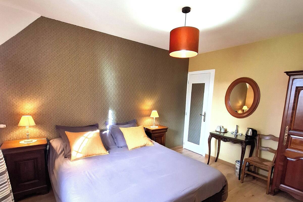 Quarto com uma cama grande, duas mesas de cabeceira com luminárias, cômoda de madeira, uma mesinha com espelho e uma porta que dá para outro cômodo.