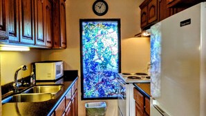 A compact kitchen with wooden cabinets, a microwave, a refrigerator, a stove, and a stained glass window. - Landfall Towers 34 Minium 1 Bedroom Condo by RedAwning (South Padre Island)