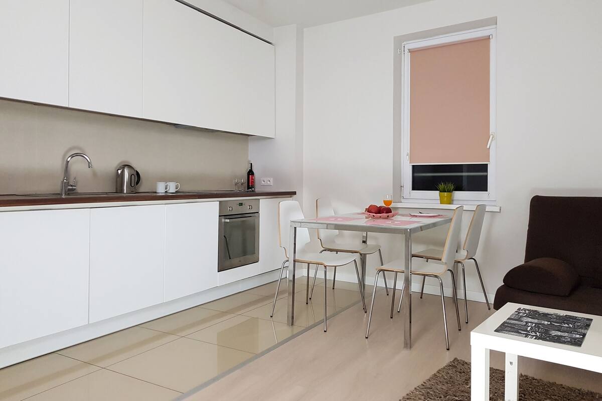 A compact kitchen with white cabinets, a stainless steel sink, and a built-in oven. There is a dining area with a table and chairs, and a brown sofa in the living space.