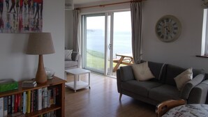 A living room with a sofa, a coffee table, a bookshelf, a lamp, a painting, a clock, and a view of the sea through a glass door.